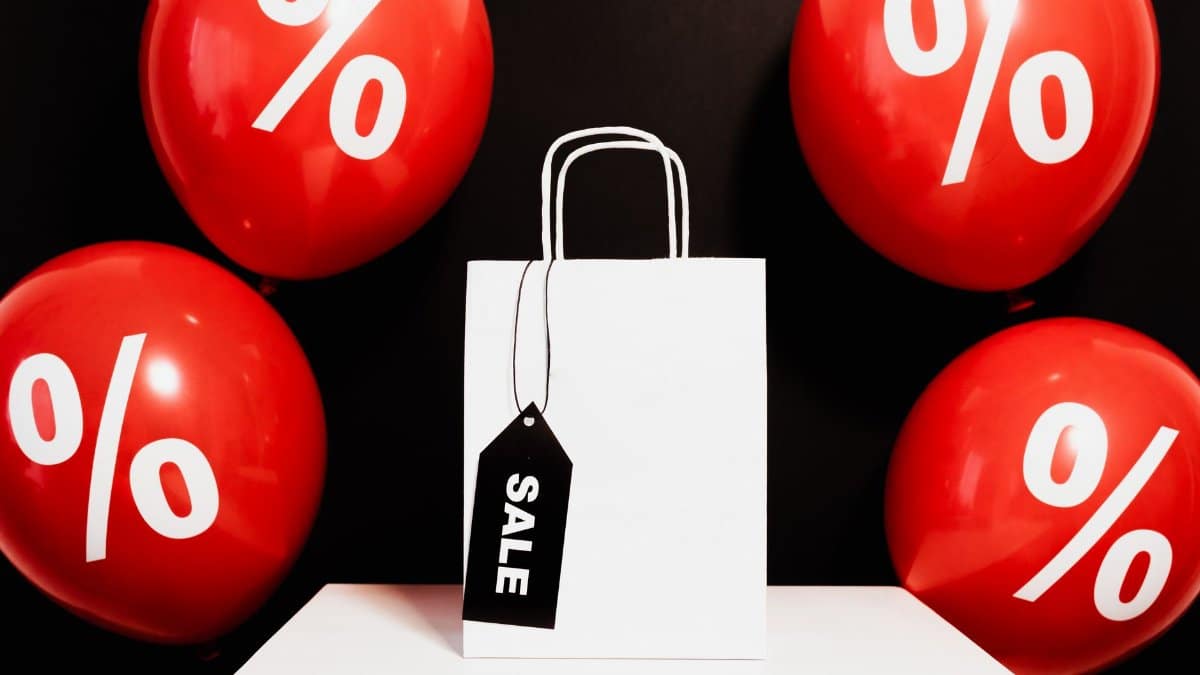 White shopping bag with sale tag surrounded by red balloons displaying percentage signs.