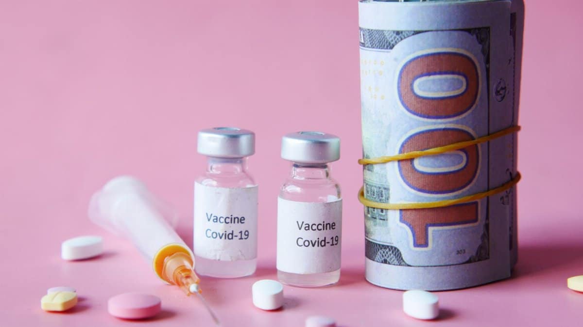Covid-19 vaccine vials, US currency, and pills on a pink background, representing healthcare costs.