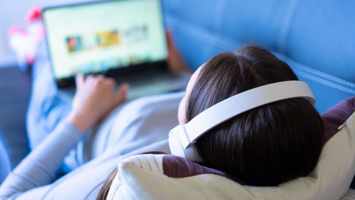 A woman lying on a sofa, enjoying music with headphones and browsing the internet on a laptop.