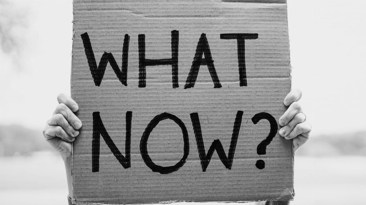 A close-up of hands holding a cardboard sign with the text 'What Now?'