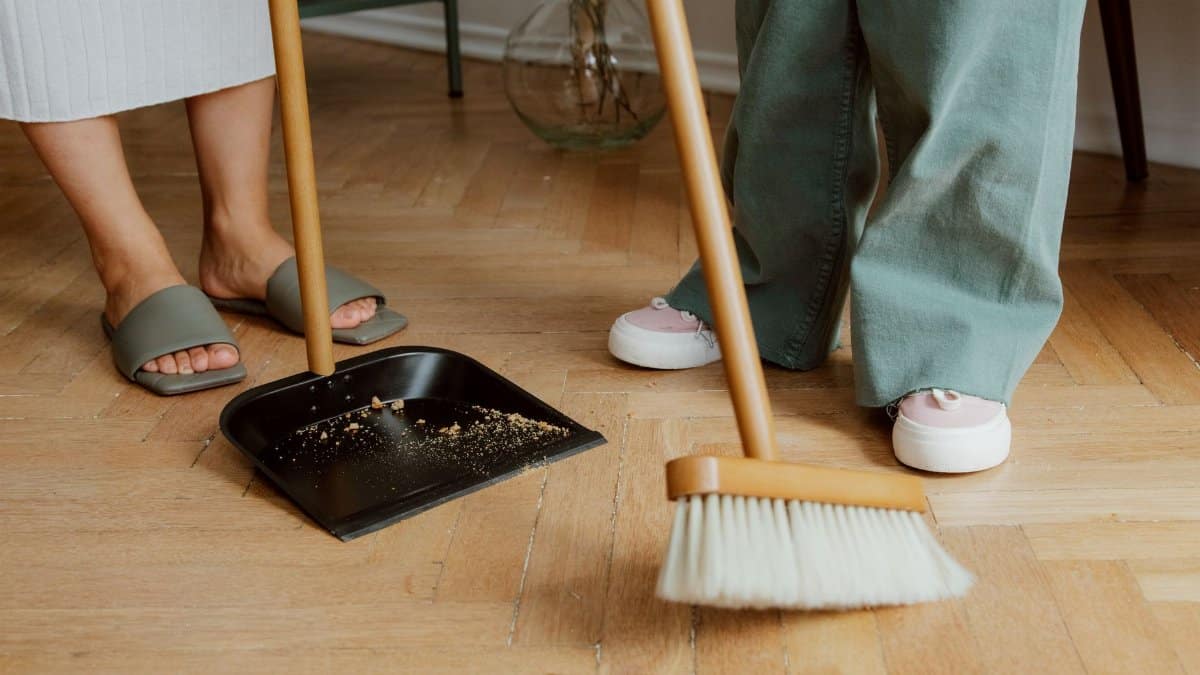Close-up of people cleaning wooden floor with broom and dustpan indoors.