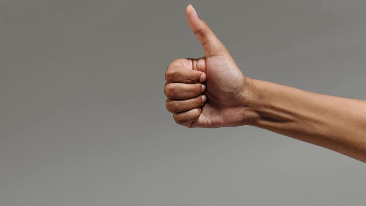 A close-up image of a hand making a thumbs up gesture against a neutral background.
