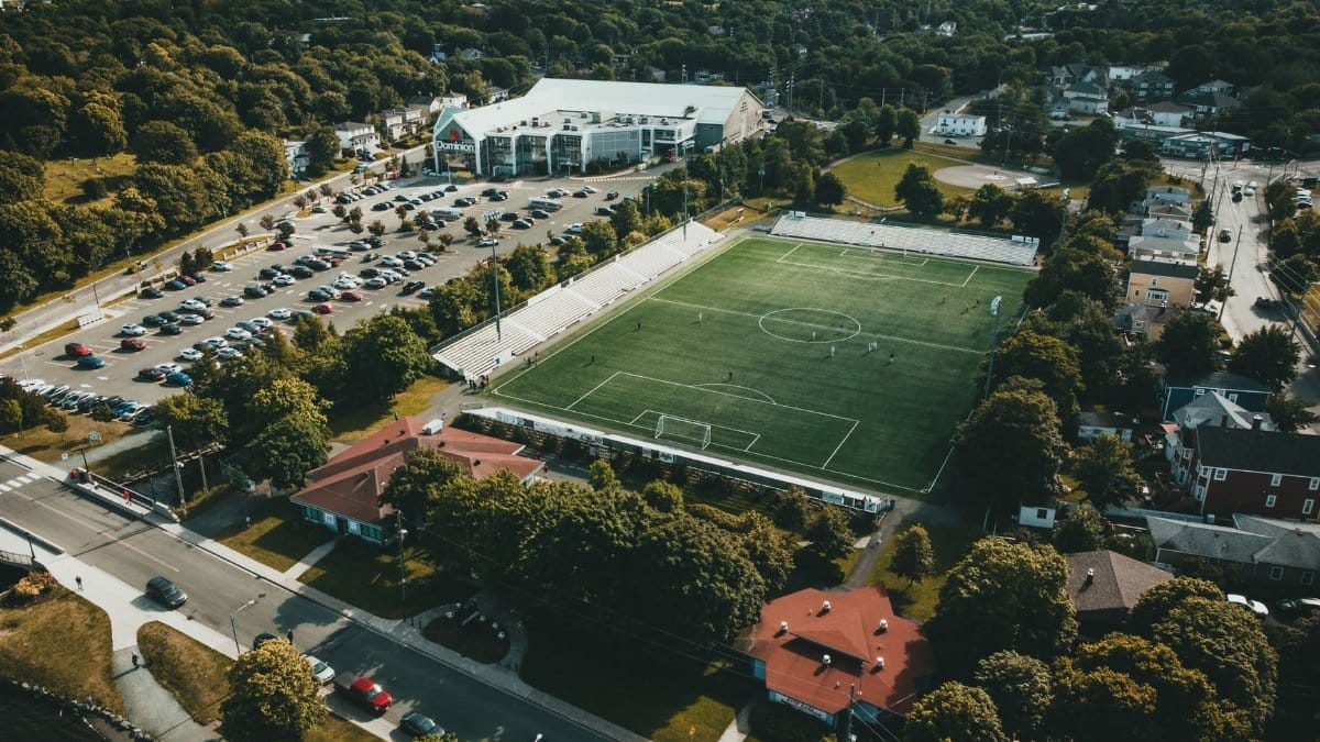 Aerial shot showcasing a soccer field in a vibrant town setting, surrounded by parking and greenery.