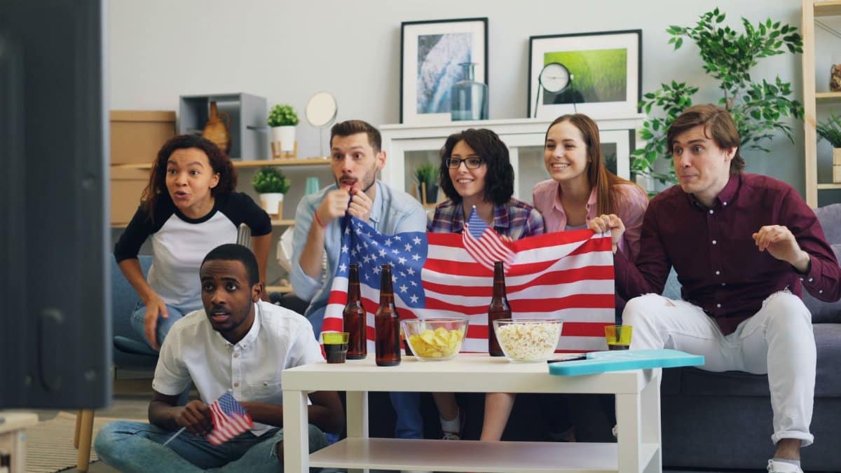 A lively group of friends celebrates while watching sports indoors, holding American flags and enjoying snacks.