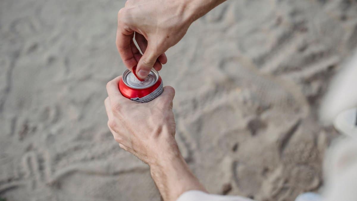 From above of crop anonymous male opening can of drink standing on sandy beach