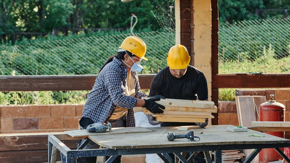 Two construction workers organize wooden planks on a table at a sunny outdoor site.