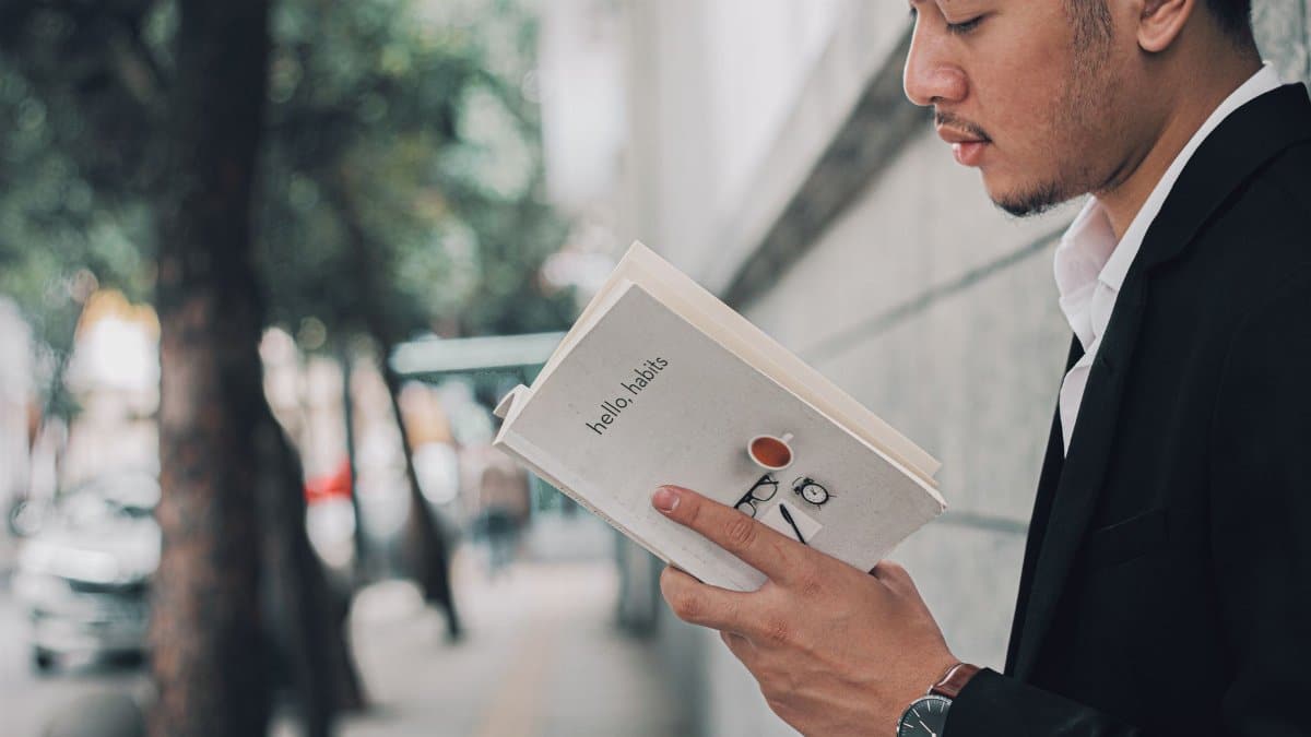 A man in a suit reading a book titled 'hello habits' on a city sidewalk.