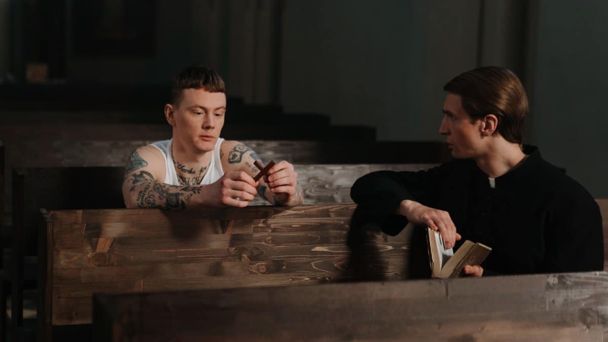 A priest and a tattooed man have a thoughtful conversation in a church setting, holding a cross and a Bible.