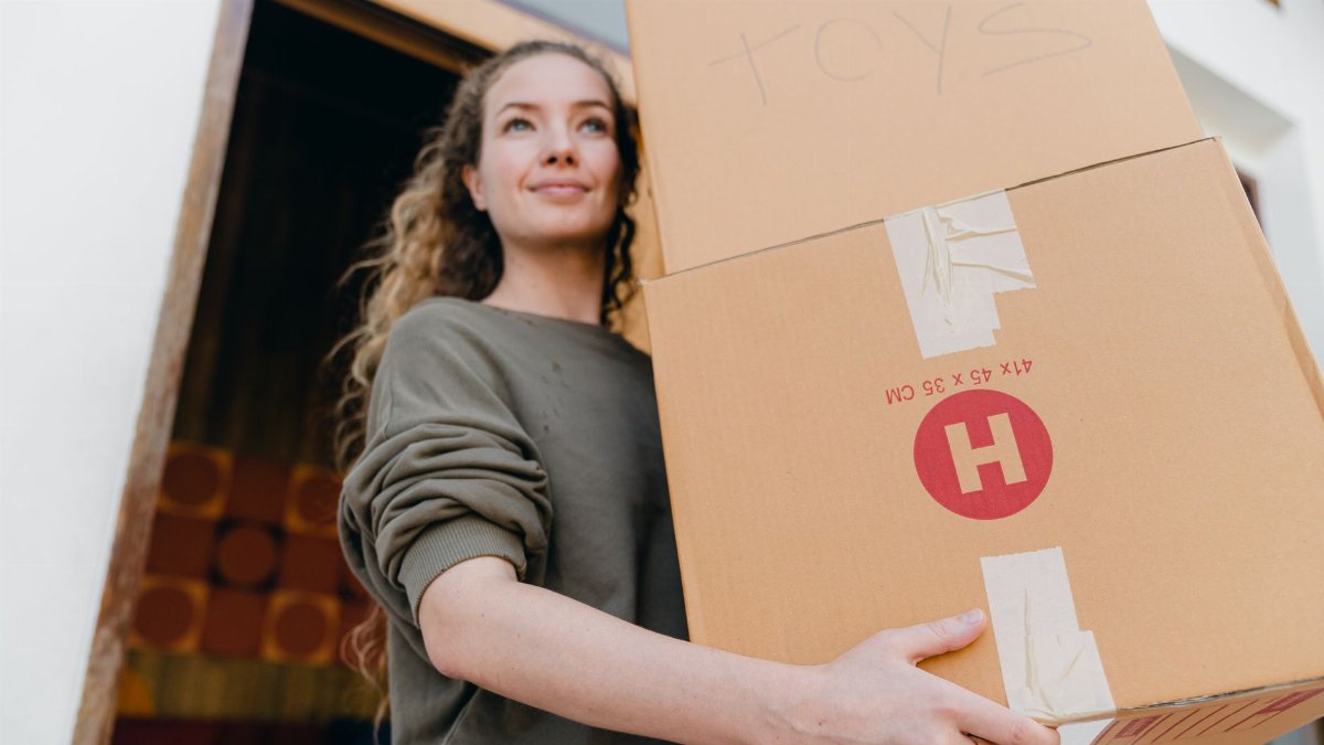 Confident young lady in casual wear carrying heavy carton boxes with toys and looking away while leaving old house