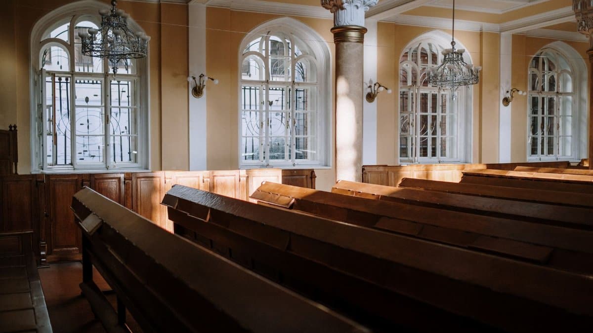 Sunlit church interior with arched windows, wooden pews, and chandeliers.