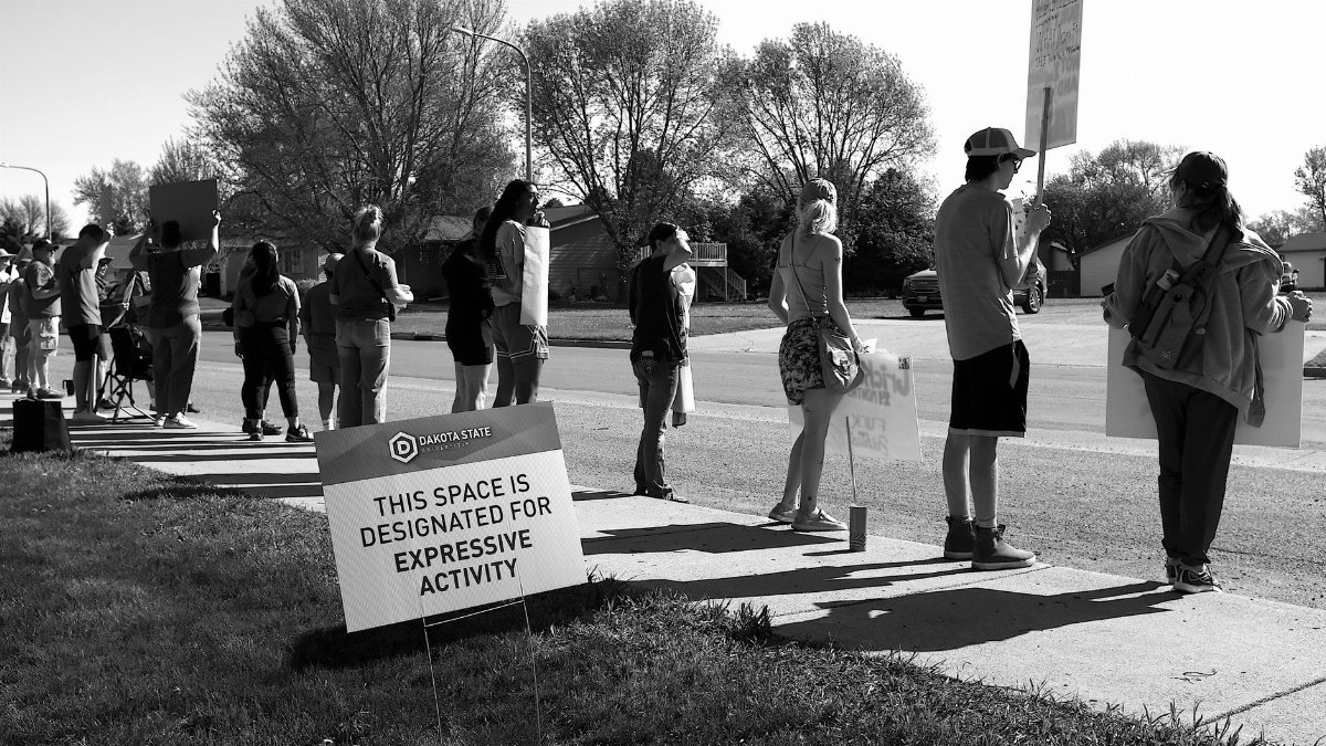 A group of people holding signs in a designated expressive activity zone during a peaceful protest outdoors.