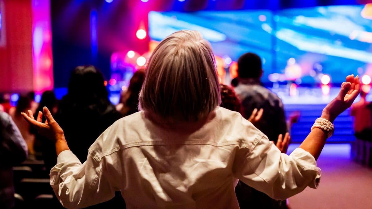 A vibrant indoor worship service with people raising hands in praise and colorful stage lighting.