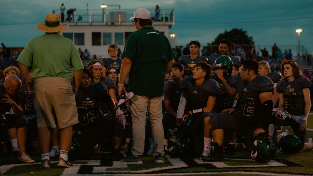 Football team huddle with coaches during an evening game at an outdoor stadium.