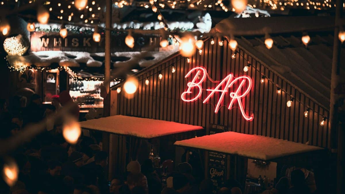 A cozy outdoor bar with a vibrant neon sign, surrounded by warm holiday lights at night.