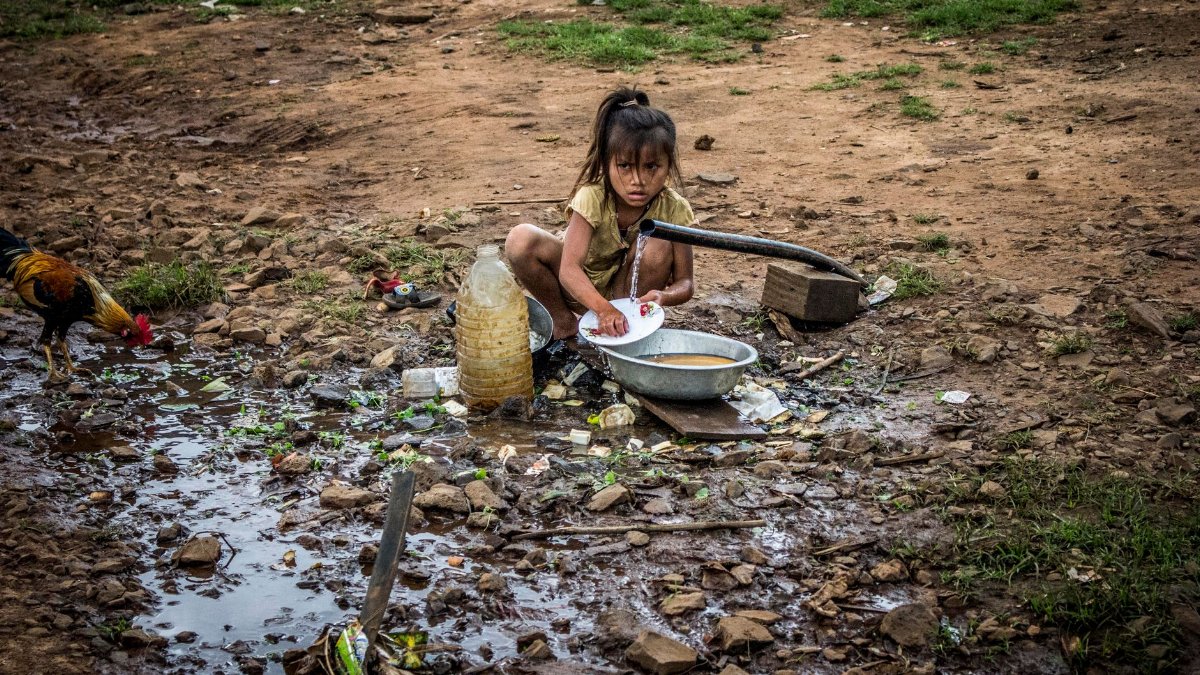 A young girl washes dishes at an outdoor hand pump, reflecting rural daily life.