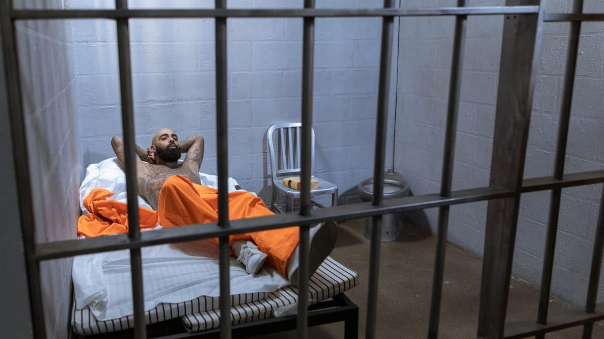 A tattooed inmate in an orange uniform lies on a bed inside a prison cell.