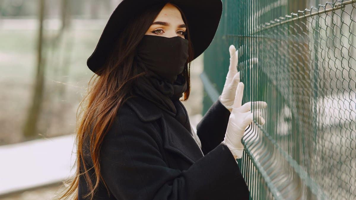 Side view of young unrecognizable female in outerwear and medical mask and gloves standing near fence and looking through holes while walking around city park