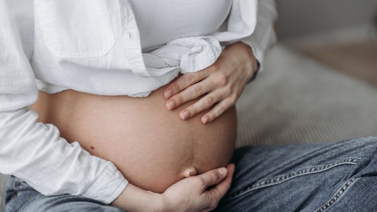 Close-up of a pregnant woman sitting, cradling her bare belly with both hands.