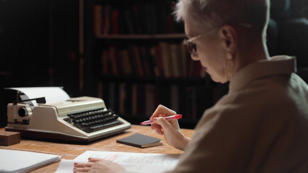 Elderly woman reviewing a manuscript with a typewriter at a wooden desk.