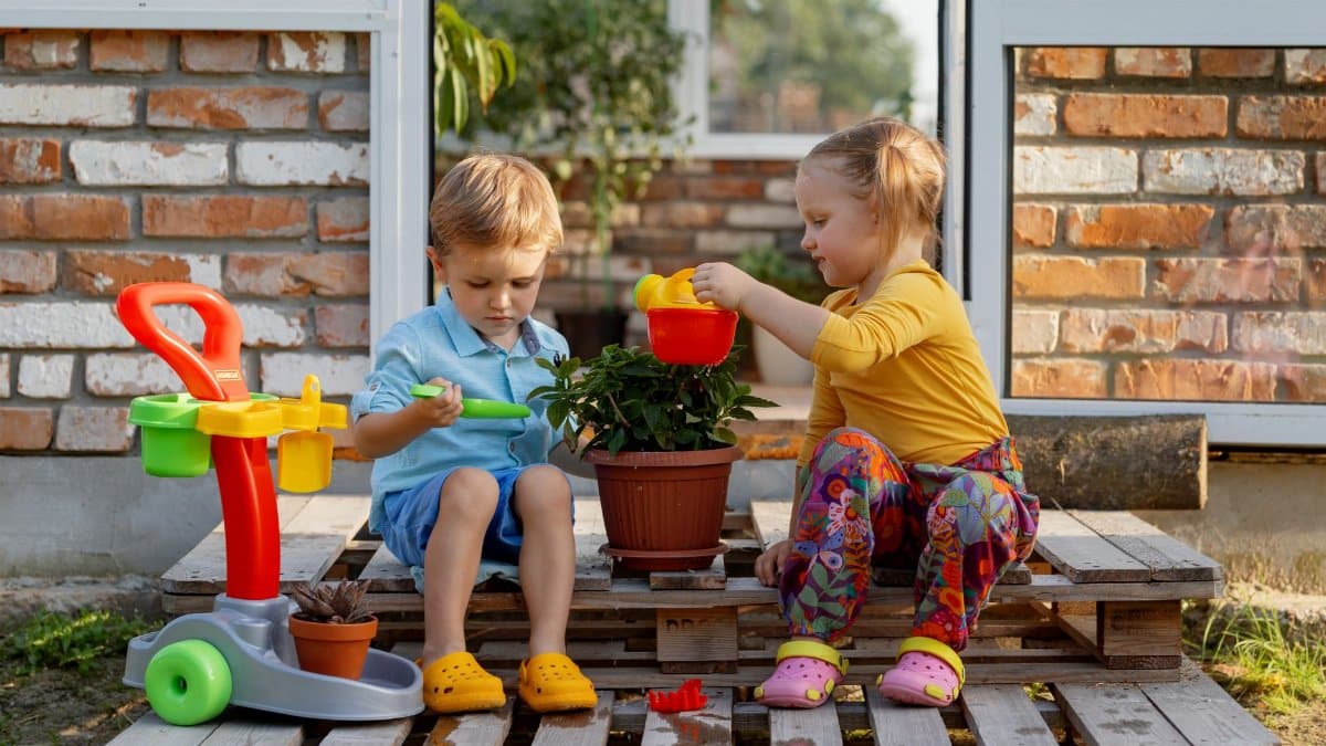 Two children enjoying gardening and playtime with toys outside a greenhouse.