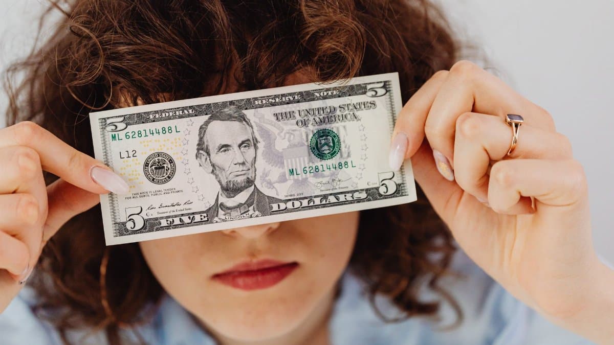 Woman with curly hair playfully covering eyes with a five dollar bill.