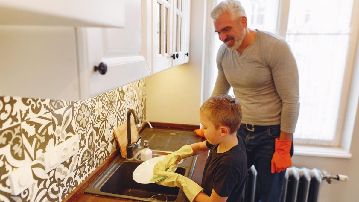 From above of grey haired father with beard teaching son dishwashing at kitchen