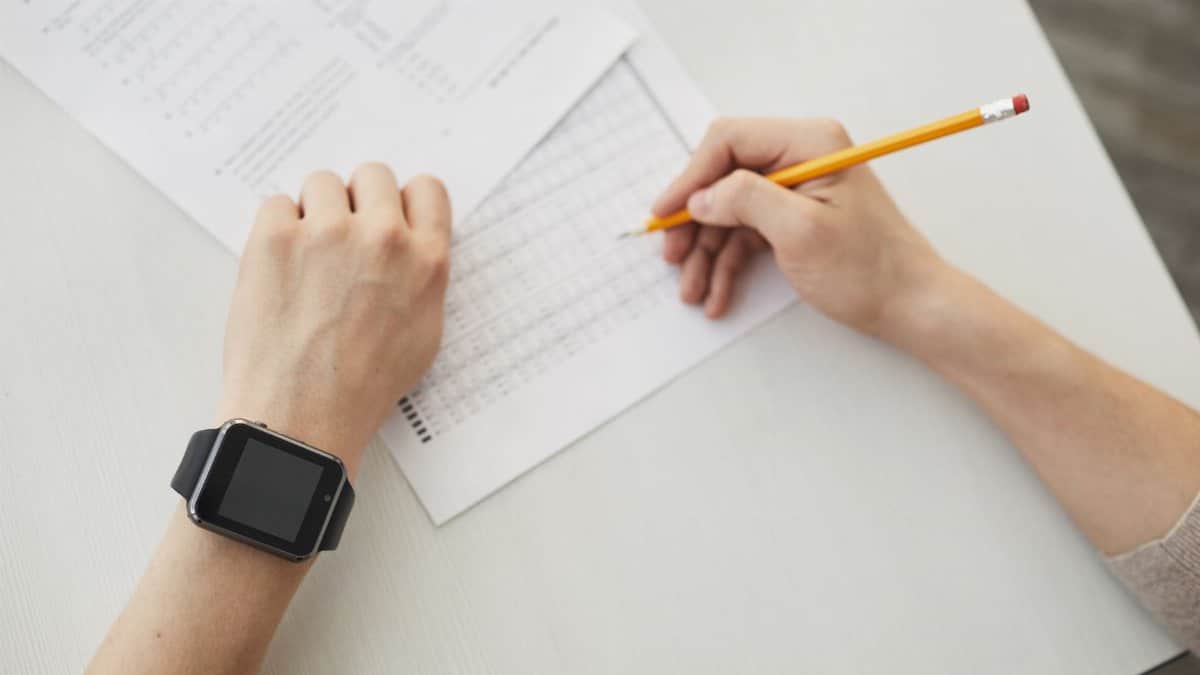 Unrecognizable student writing an exam using a pencil, watch visible on wrist.