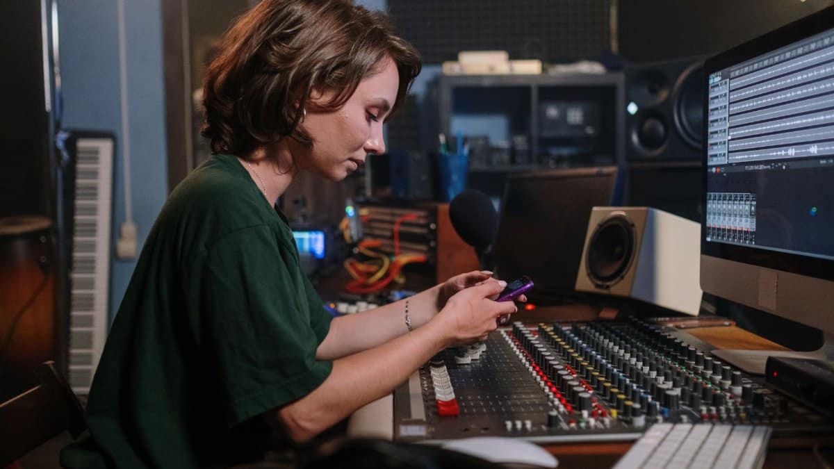 A woman in a green t-shirt uses her smartphone while sitting at a sound mixer in a recording studio.
