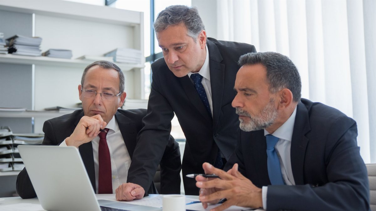 Three businessmen in suits collaborating in a modern office setting, focused on a laptop.