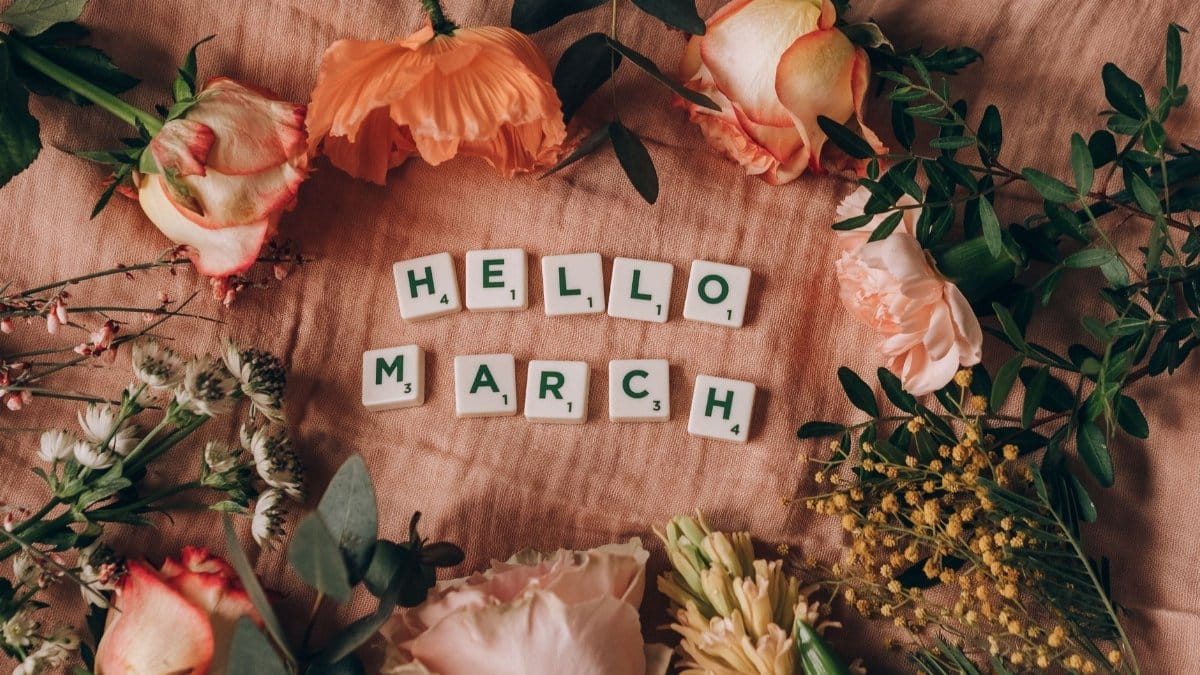 Flat lay floral arrangement with Scrabble tiles spelling 'Hello March' on a pink surface.