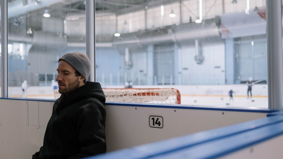 A man wearing a knit cap leans against the wall inside an indoor ice hockey rink.