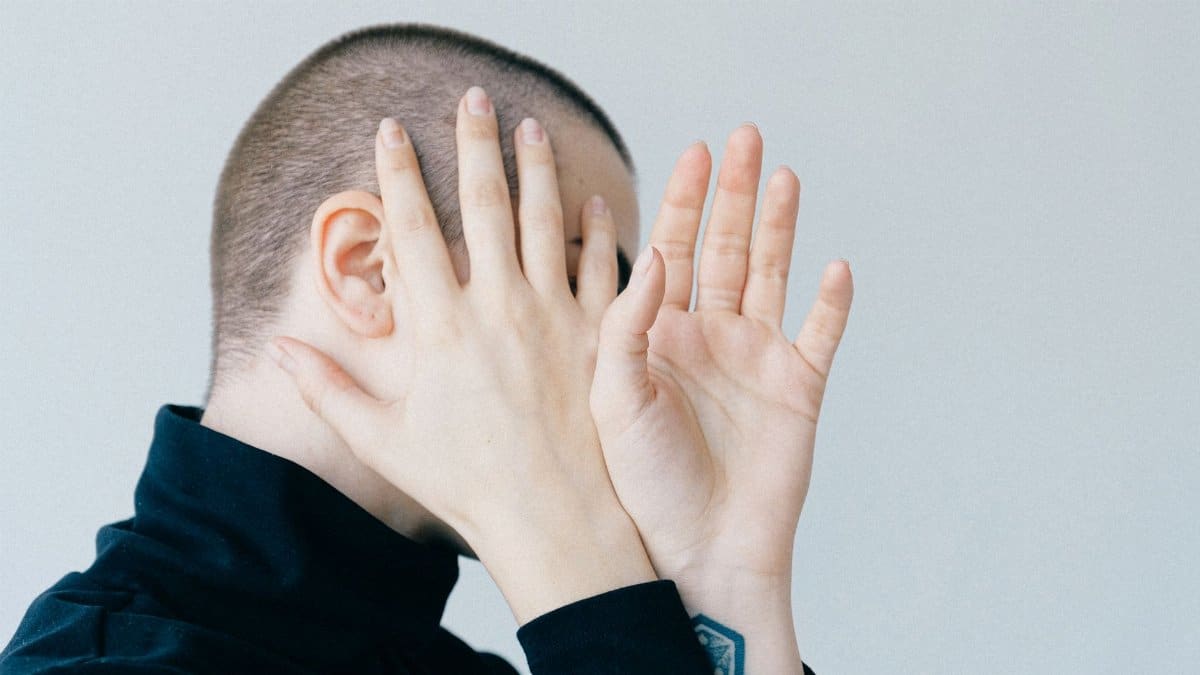 A person hides their face with hands in a minimalist portrait with a light background.