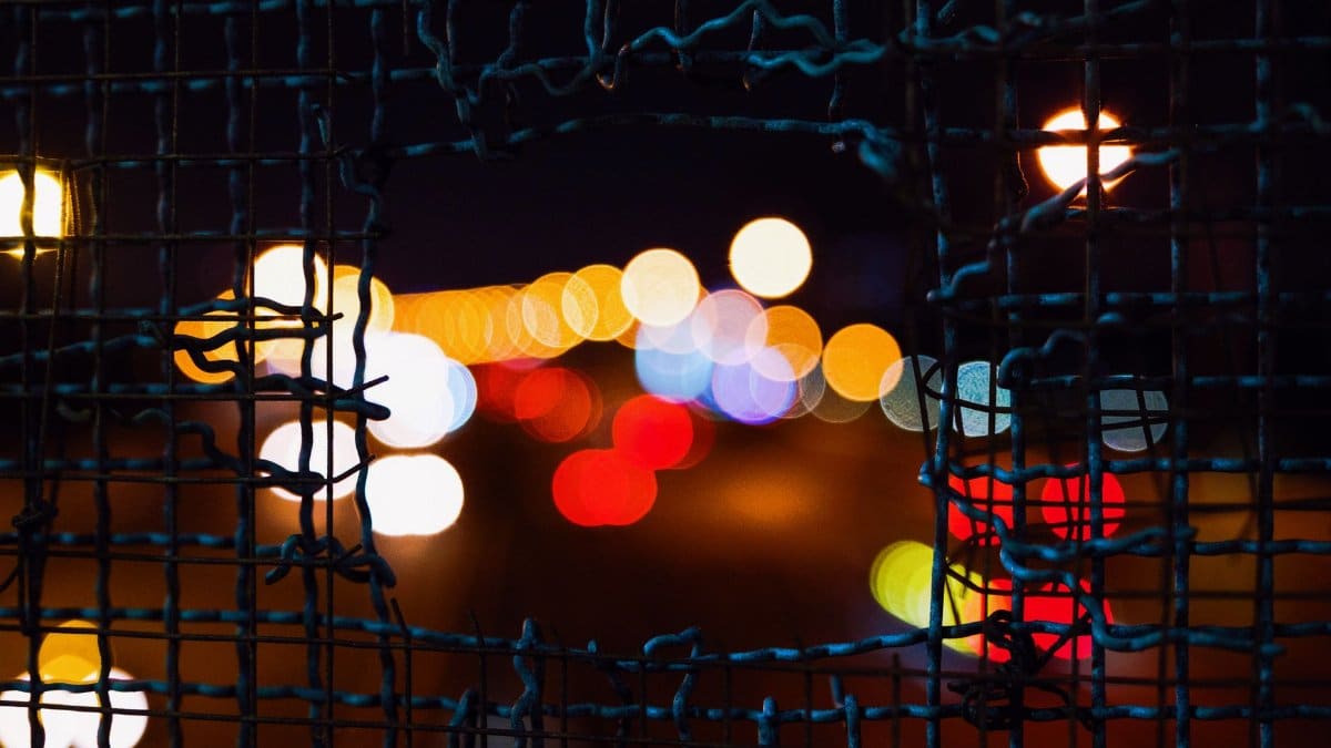 A close-up of a broken metal fence showcasing colorful bokeh lights at night.