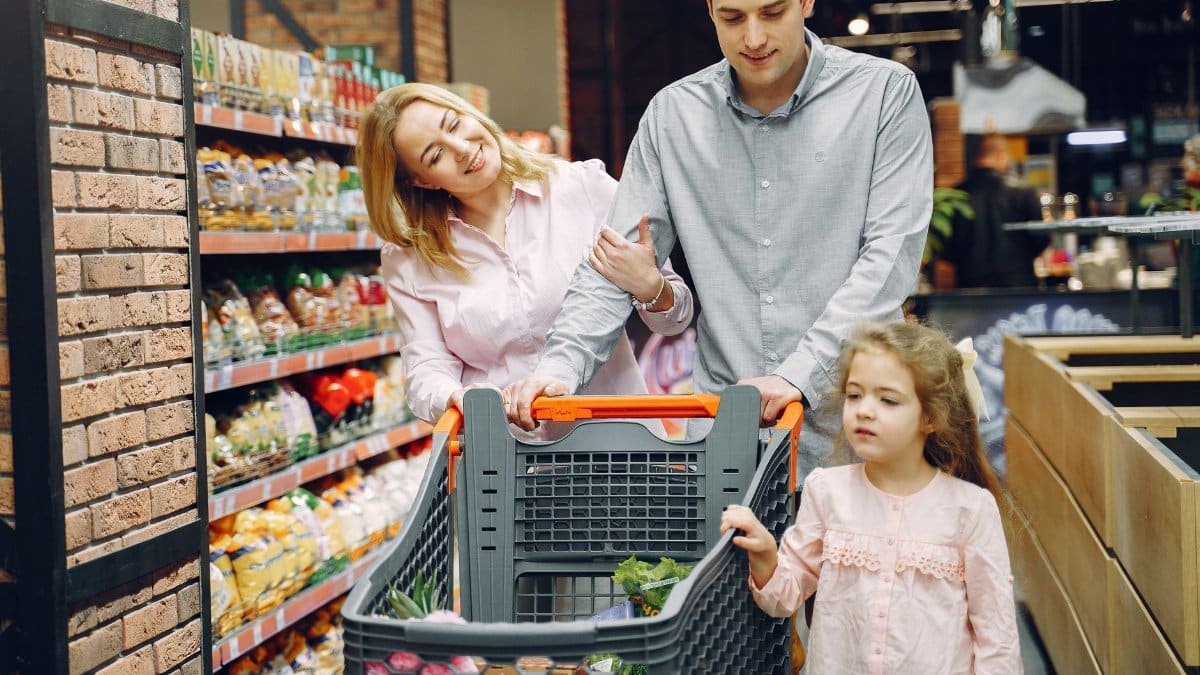 A family of three shopping in a supermarket, pushing a cart full of groceries.