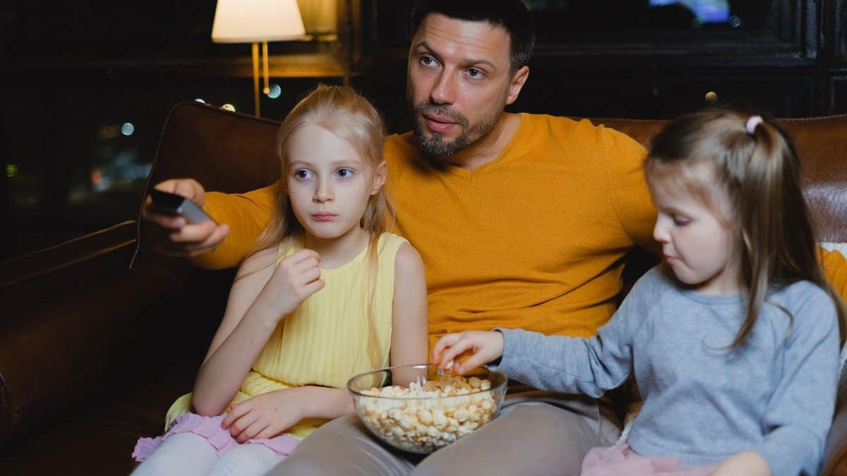 A father and his two daughters enjoying a movie night with popcorn on the couch.