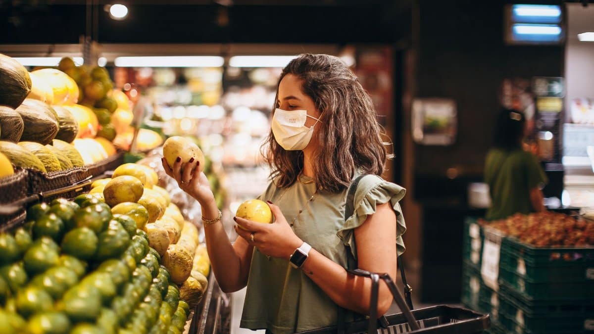 A woman wearing a mask selects fresh fruit in a grocery store aisle.