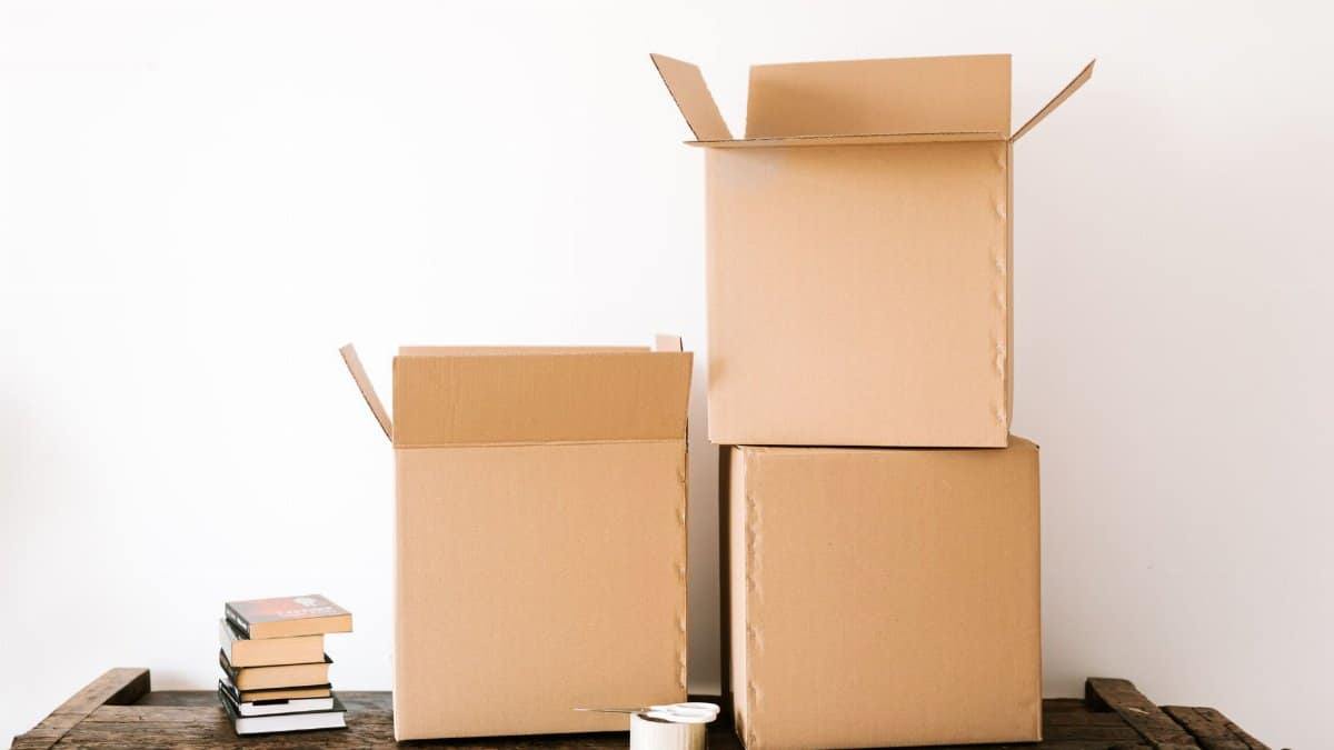 Opened cardboard boxes and stack of books placed on wooden shabby table near tape with scissors against white plain wall