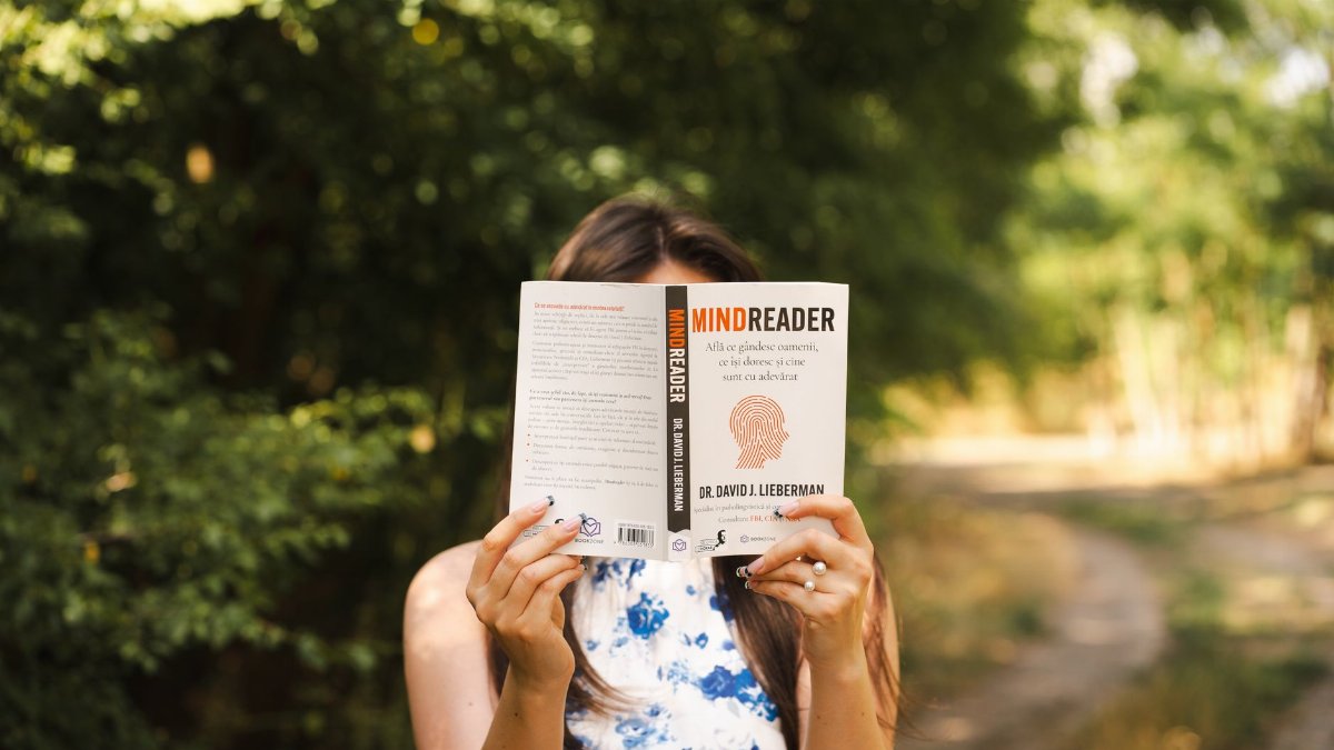 A woman enjoys reading a book while standing in a sunny forest, surrounded by lush greenery.