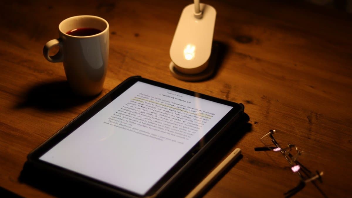 A tablet with a document, coffee, and glasses on a wooden desk in warm evening lighting.
