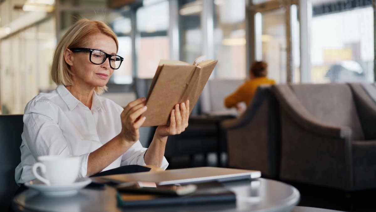 Elegant woman reading a book at a cafe table, enjoying leisure time with coffee nearby.