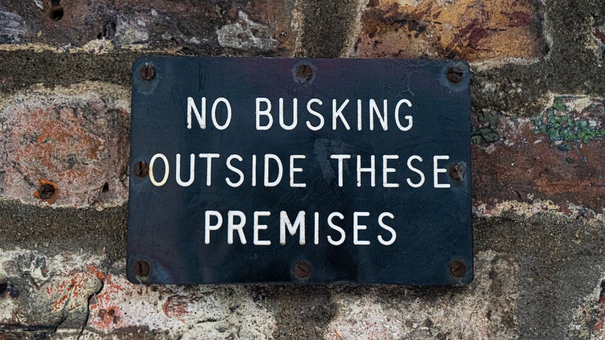 Black sign stating 'No Busking' affixed to a textured brick wall surface.
