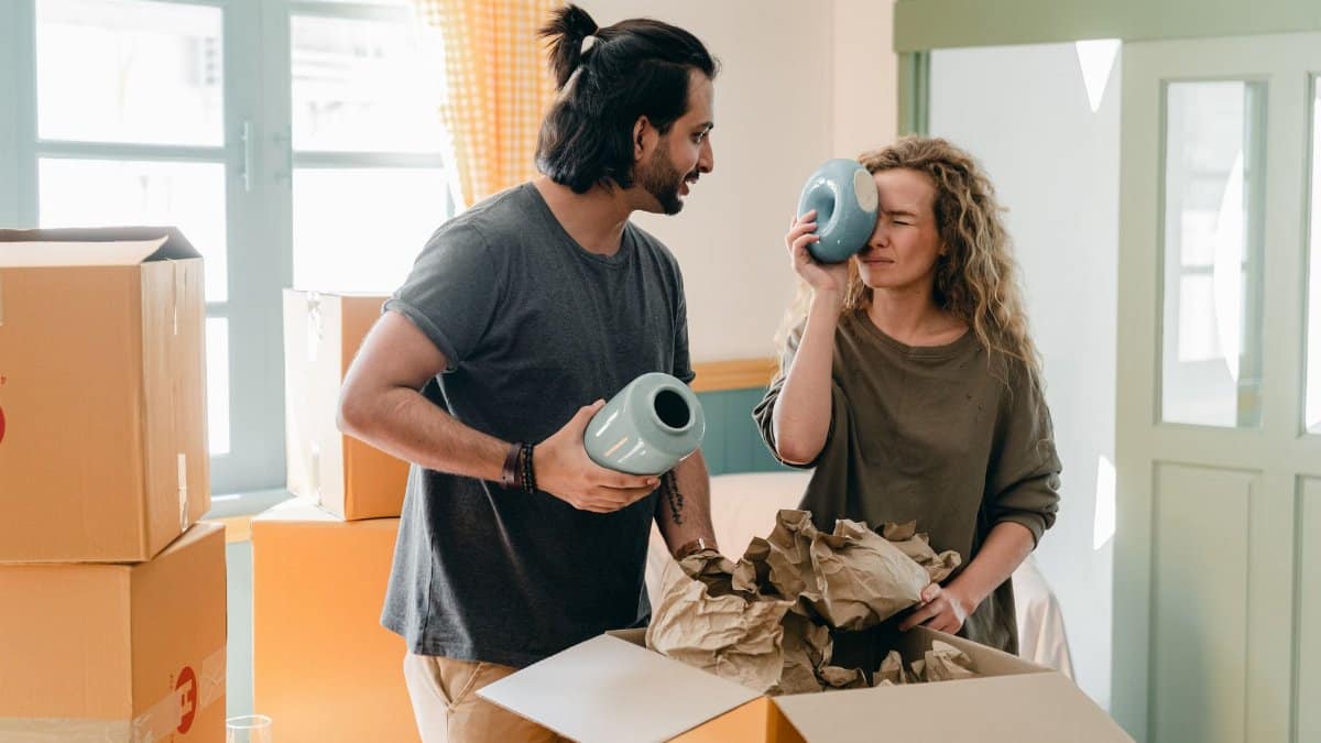 Cheerful ethnic man in smart watch with blue ceramic vase interacting with funny girlfriend with closed eyes and big souvenir standing near heap of cardboard boxes preparing to relocate