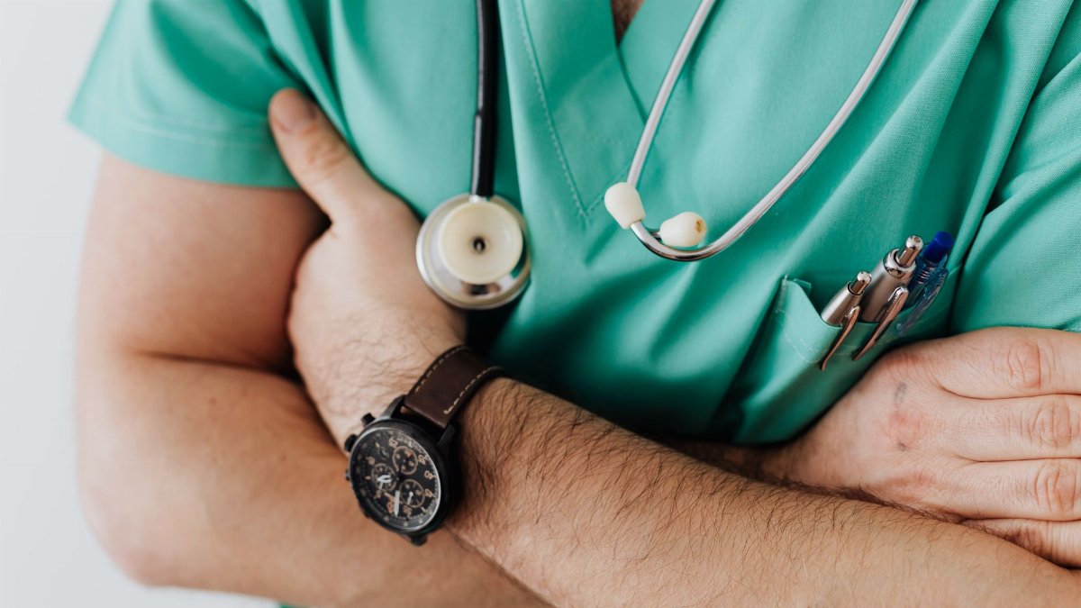 Unrecognizable man wearing medical uniform with stethoscope and pens standing in hospital with crossed arms during work in day time