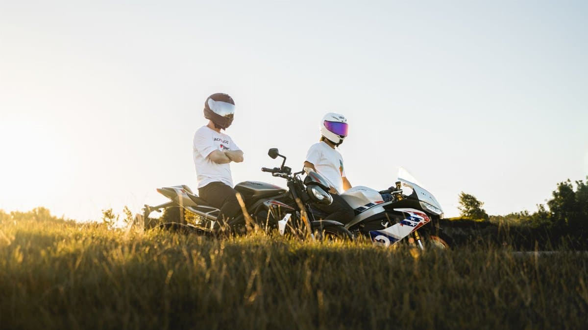 Two motorcyclists in helmets pose with their bikes in a grassy countryside at sunset.