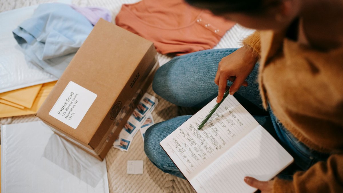 Top view of crop anonymous female checking address on parcel and in planner among clothes on floor