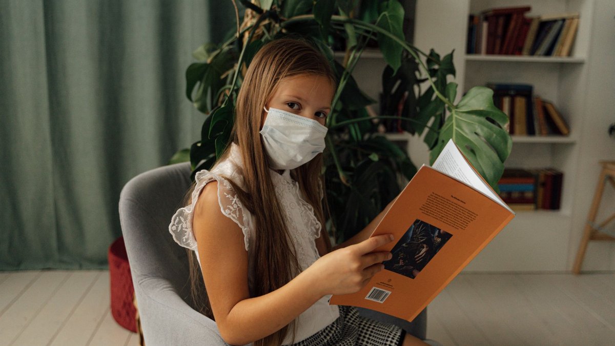 A girl sits indoors, reading a book while wearing a face mask, embracing the new normal.