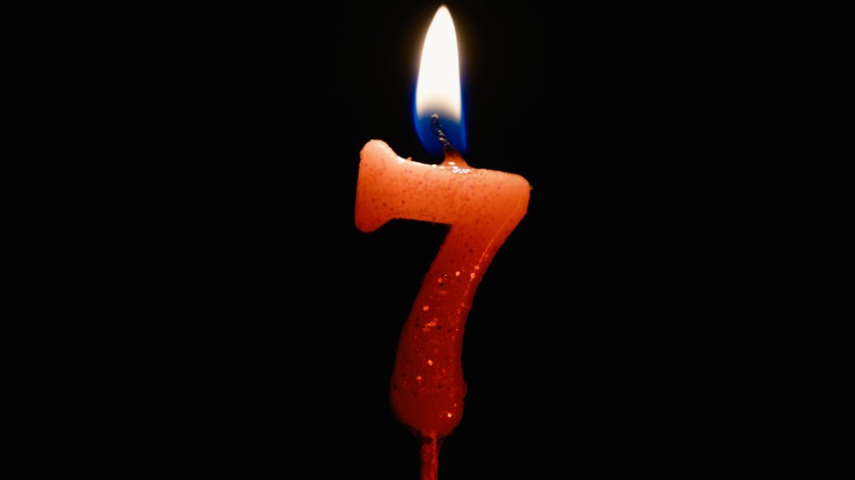 An image of a lit number seven candle with a vibrant flame against a dark backdrop.