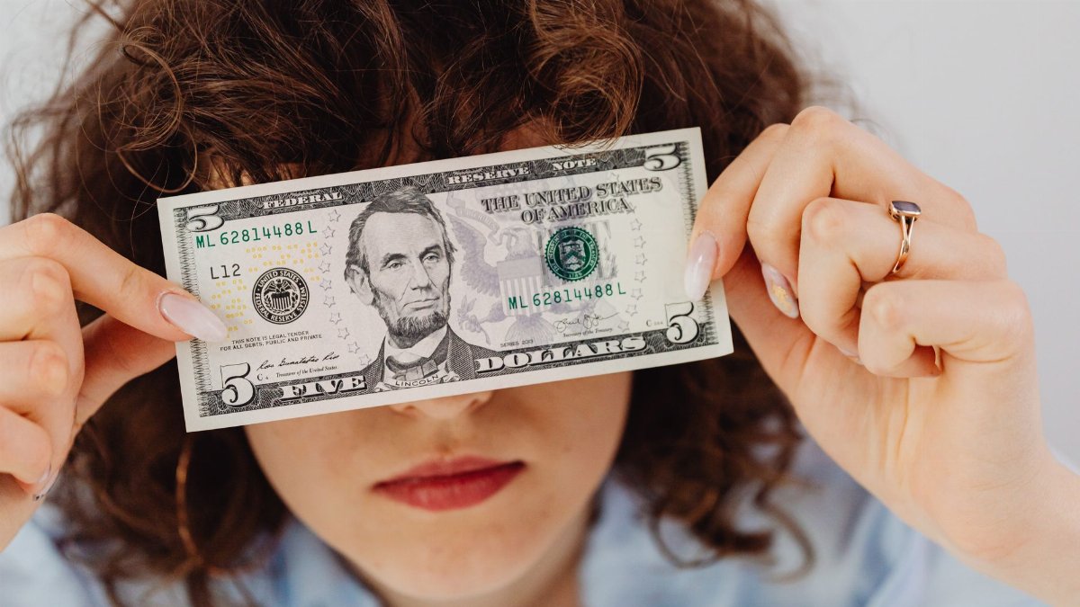 Woman with curly hair playfully covering eyes with a five dollar bill.