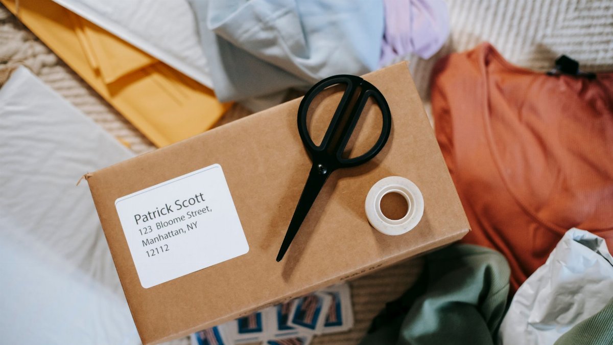 Top view of tape with scissors on carton box with exact address and name among clothes