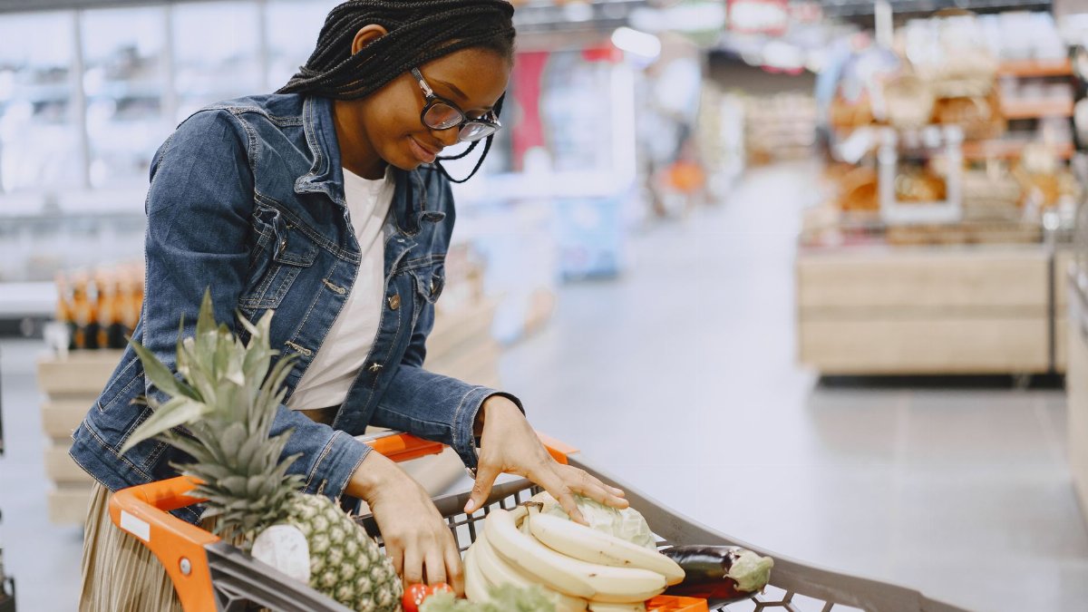 A woman shops for fresh fruits and vegetables in a supermarket, pushing a cart filled with groceries.
